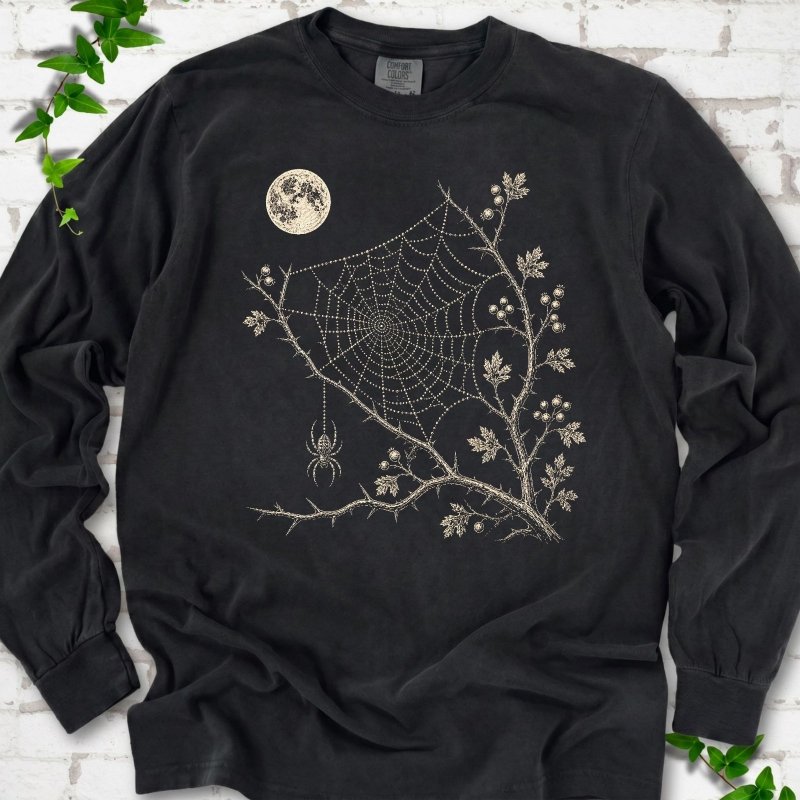 Black Comfort Colors® long sleeve T-shirt flat lay featuring a cream moon, spider web, berry branches and hanging spider design.