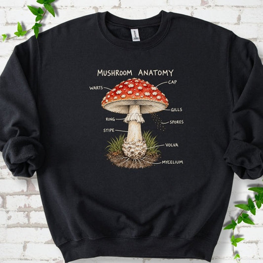 Black crewneck sweatshirt with a red-capped fly agaric mushroom diagram and ivory hand-lettered labels, flat lay on white brick with ivy