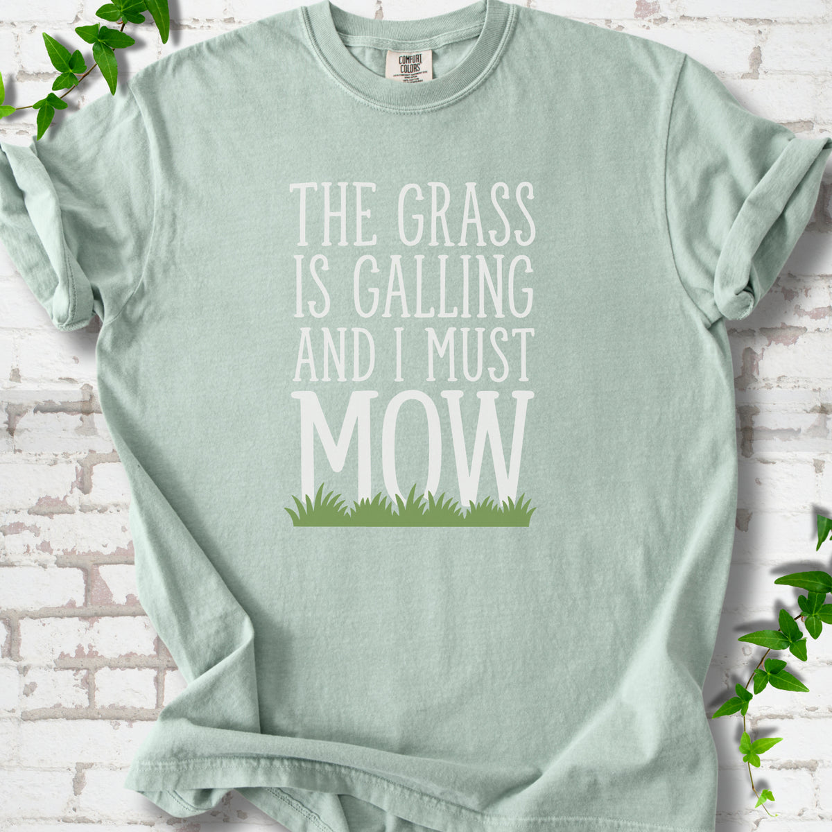 Bay short sleeve T-shirt flat lay with cream "The grass is calling and I must mow" lettering above a green grass silhouette, on white brick with ivy sprigs.