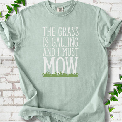 Bay short sleeve T-shirt flat lay with cream "The grass is calling and I must mow" lettering above a green grass silhouette, on white brick with ivy sprigs.