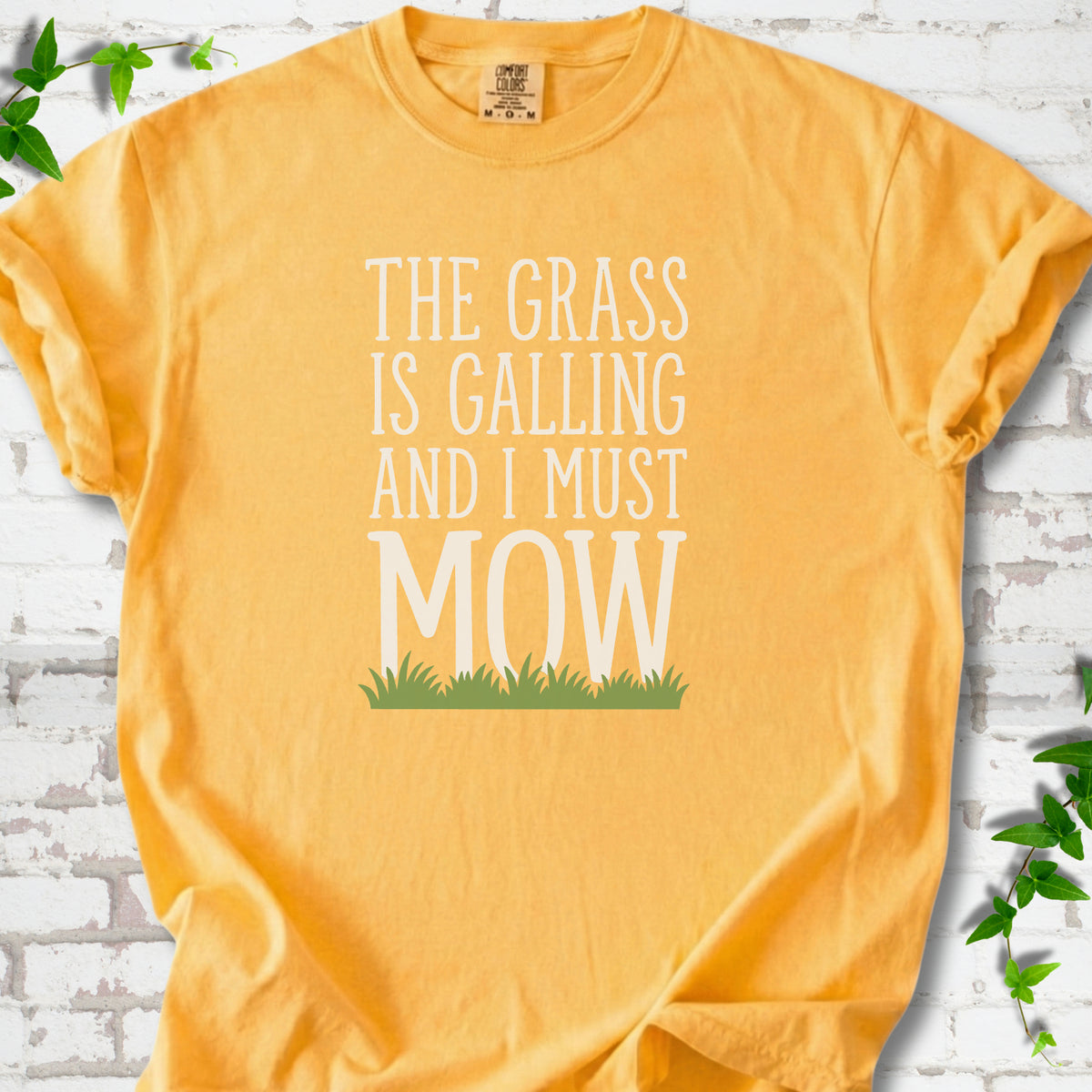 Citrus short sleeve T-shirt flat lay with cream "The grass is calling and I must mow" lettering above a green grass silhouette, on white brick with ivy sprigs.