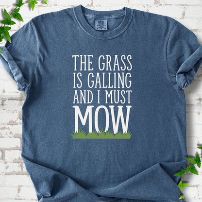 Denim short sleeve T-shirt flat lay with cream "The grass is calling and I must mow" lettering above a green grass silhouette, on white brick with ivy sprigs.