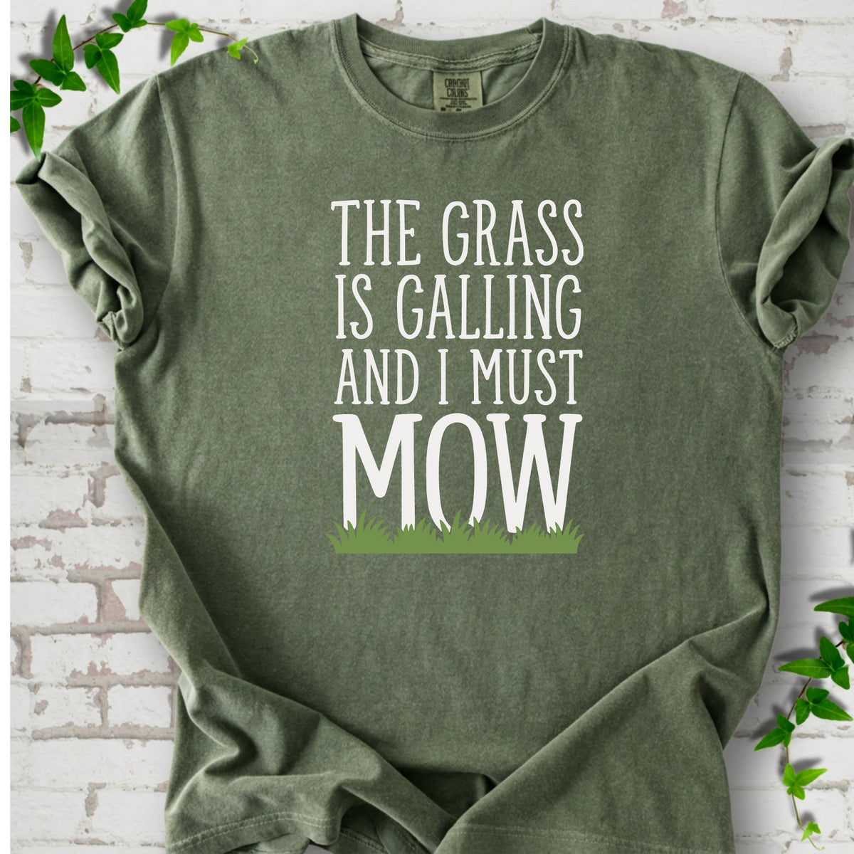 Moss short sleeve T-shirt flat lay with cream "The grass is calling and I must mow" lettering above a green grass silhouette, on white brick with ivy sprigs.