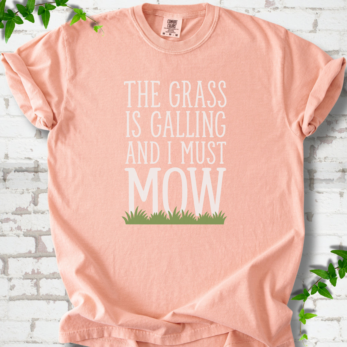 Peachy short sleeve T-shirt flat lay with cream "The grass is calling and I must mow" lettering above a green grass silhouette, on white brick with ivy sprigs.