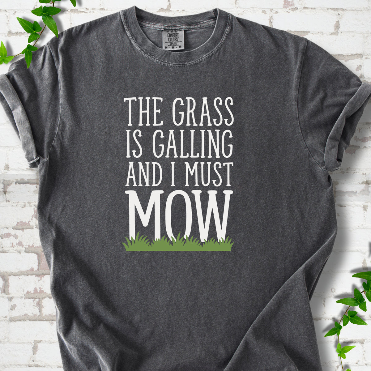Pepper short sleeve T-shirt flat lay with cream "The grass is calling and I must mow" lettering above a green grass silhouette, on white brick with ivy sprigs.