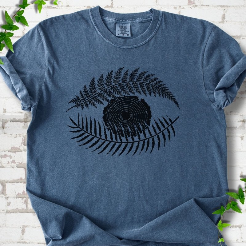 Denim Comfort Colors® T-shirt flat lay featuring a black botanical eye formed by fern fronds and a tree-ring centre.