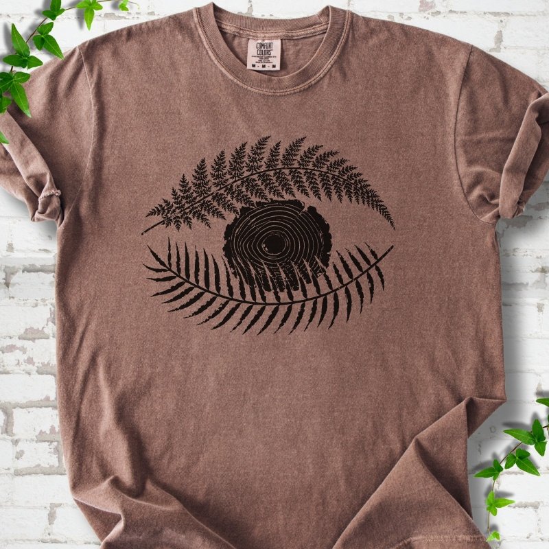 Espresso Comfort Colors® T-shirt flat lay featuring a black botanical eye formed by fern fronds and a tree-ring centre.