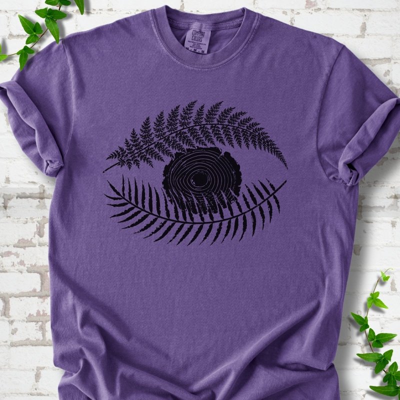 Grape Comfort Colors® T-shirt flat lay featuring a black botanical eye formed by fern fronds and a tree-ring centre.