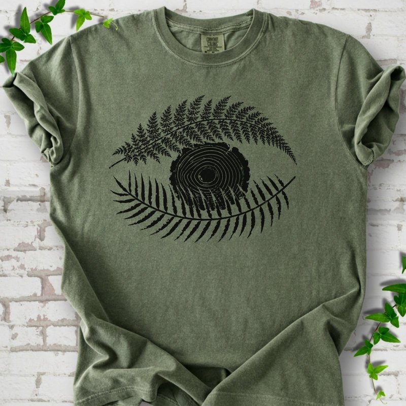 Moss Comfort Colors® T-shirt flat lay featuring a black botanical eye formed by fern fronds and a tree-ring centre.