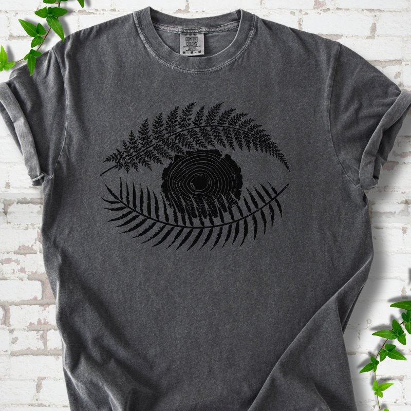Pepper Comfort Colors® T-shirt flat lay featuring a black botanical eye formed by fern fronds and a tree-ring centre.