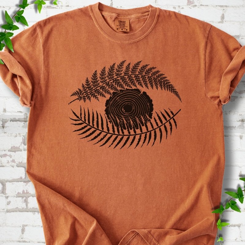 Yam Comfort Colors® T-shirt flat lay featuring a black botanical eye formed by fern fronds and a tree-ring centre.