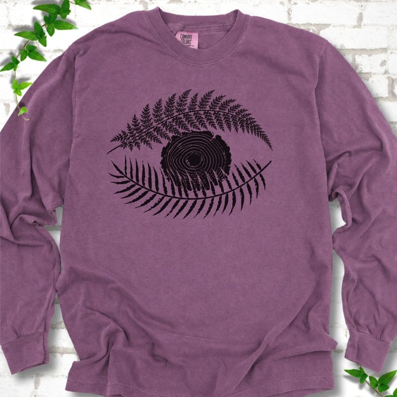 Berry Comfort Colors® long sleeve T-shirt flat lay featuring a black botanical eye formed by fern fronds and a tree-ring centre.