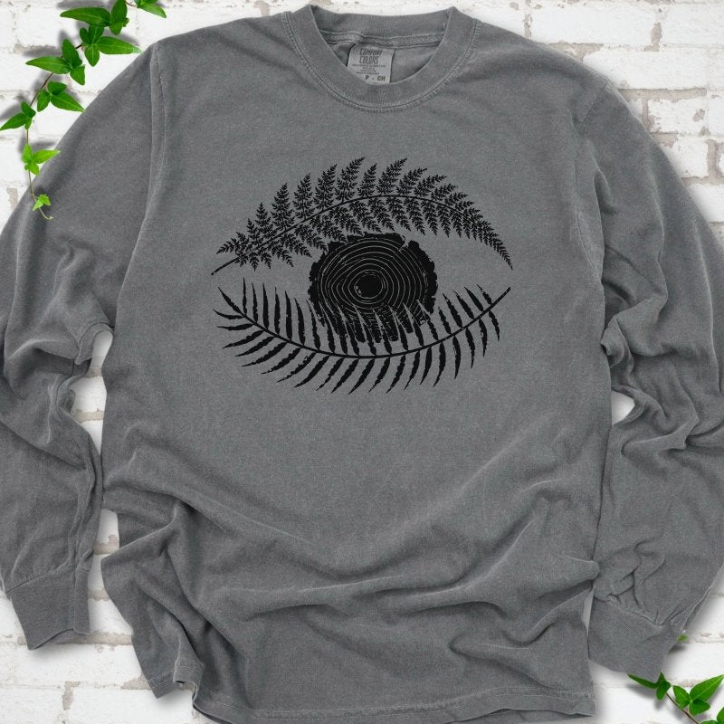 Pepper Comfort Colors® long sleeve T-shirt flat lay featuring a black botanical eye formed by fern fronds and a tree-ring centre.