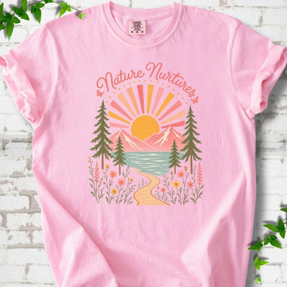 Blossom T-shirt laid flat, featuring the words 'Nature Nurtures' above a radiating sun, pink mountains, evergreens, blue-green water, a winding path and flowers.