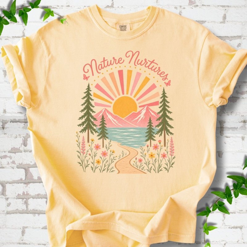 Butter T-shirt laid flat, featuring the words 'Nature Nurtures' above a radiating sun, pink mountains, evergreens, blue-green water, a winding path and flowers.