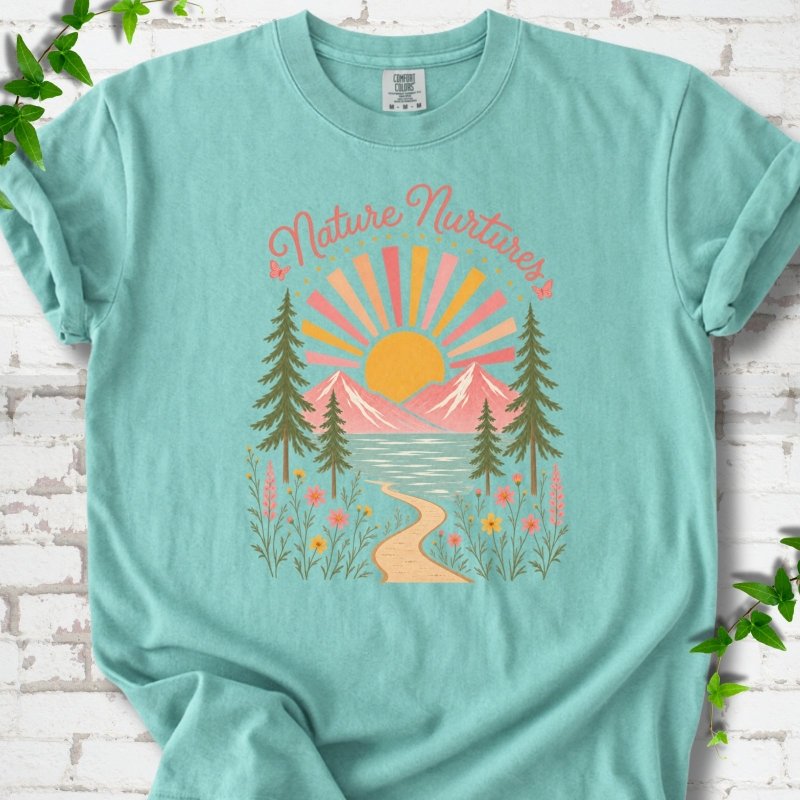 Chalky Mint T-shirt laid flat, featuring the words 'Nature Nurtures' above a radiating sun, pink mountains, evergreens, blue-green water, a winding path and flowers.