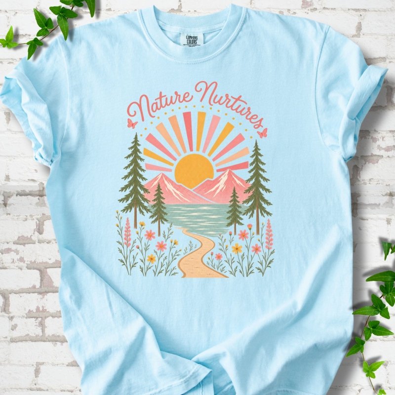 Chambray T-shirt laid flat, featuring the words 'Nature Nurtures' above a radiating sun, pink mountains, evergreens, blue-green water, a winding path and flowers.