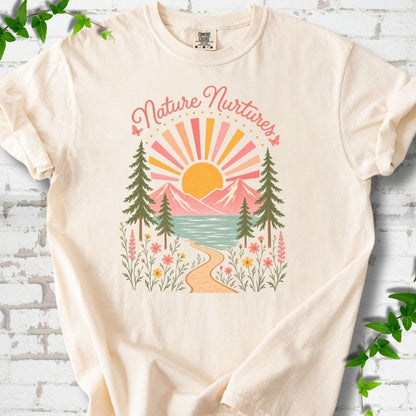Ivory T-shirt laid flat, featuring the words 'Nature Nurtures' above a radiating sun, pink mountains, evergreens, blue-green water, a winding path and flowers.