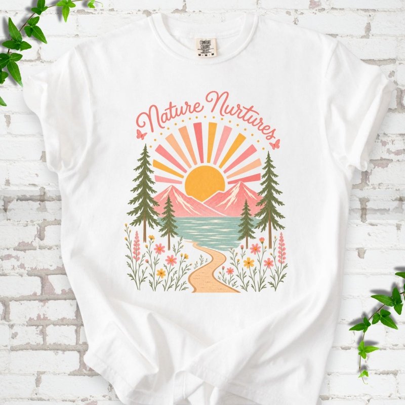 White T-shirt laid flat, featuring the words 'Nature Nurtures' above a radiating sun, pink mountains, evergreens, blue-green water, a winding path and flowers.