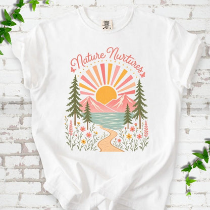 White T-shirt laid flat, featuring the words 'Nature Nurtures' above a radiating sun, pink mountains, evergreens, blue-green water, a winding path and flowers.