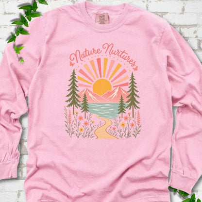 Blossom long sleeve T-shirt laid flat, featuring the words 'Nature Nurtures' above a radiating sun, pink mountains, evergreens, blue-green water, a winding path and flowers.