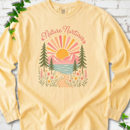 Butter long sleeve T-shirt laid flat, featuring the words 'Nature Nurtures' above a radiating sun, pink mountains, evergreens, blue-green water, a winding path and flowers.