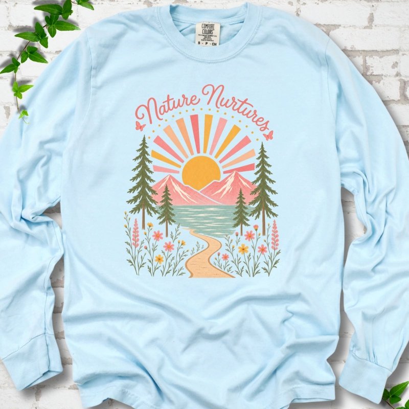 Chambray long sleeve T-shirt laid flat, featuring the words 'Nature Nurtures' above a radiating sun, pink mountains, evergreens, blue-green water, a winding path and flowers.