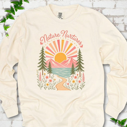 Ivory long sleeve T-shirt laid flat, featuring the words 'Nature Nurtures' above a radiating sun, pink mountains, evergreens, blue-green water, a winding path and flowers.