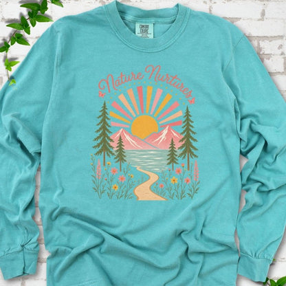 Seafoam long sleeve T-shirt laid flat, featuring the words 'Nature Nurtures' above a radiating sun, pink mountains, evergreens, blue-green water, a winding path and flowers.