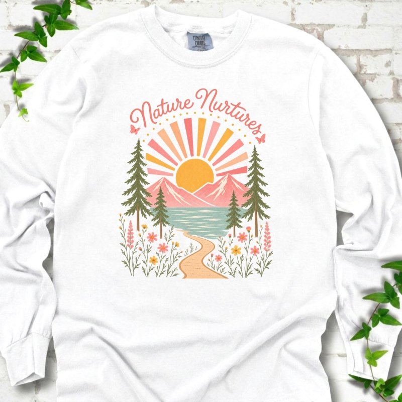 White long sleeve T-shirt laid flat, featuring the words 'Nature Nurtures' above a radiating sun, pink mountains, evergreens, blue-green water, a winding path and flowers.