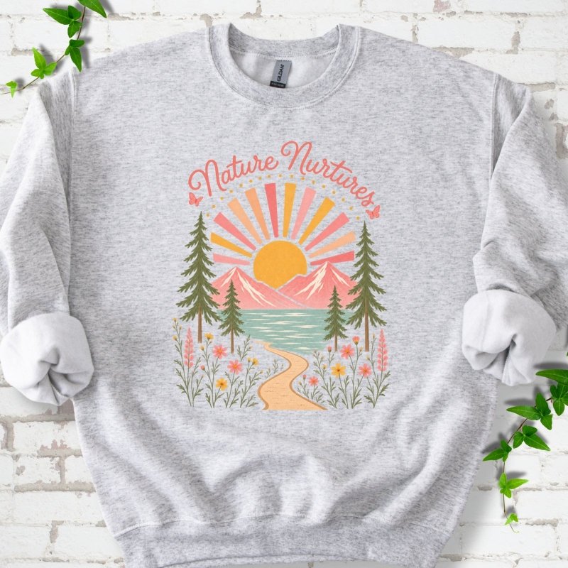 Ash sweatshirt laid flat, featuring the words 'Nature Nurtures' above a radiating sun, pink mountains, evergreens, blue-green water, a winding path and flowers.