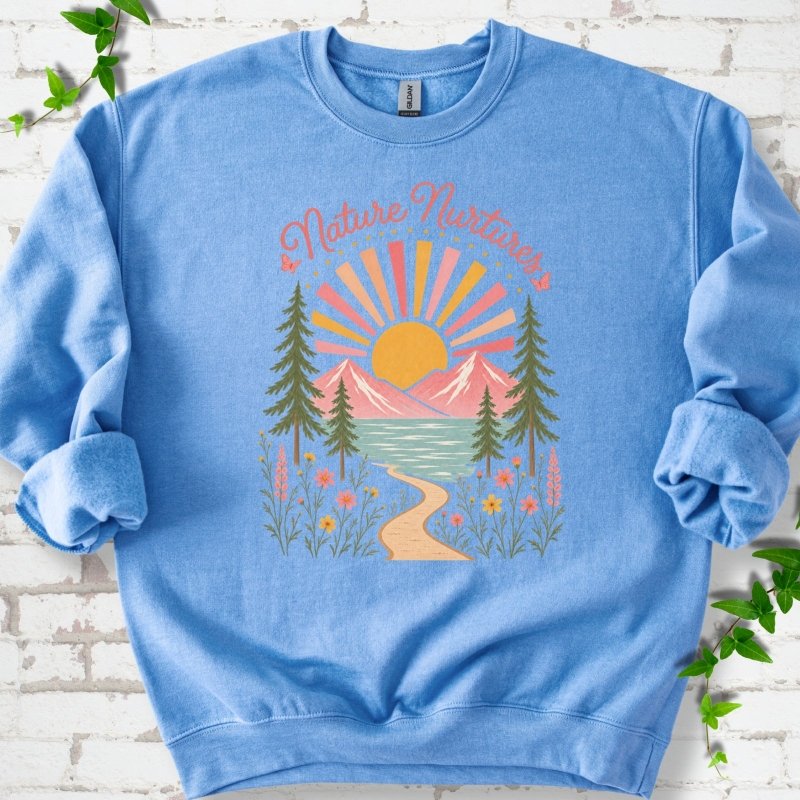 Carolina Blue sweatshirt laid flat, featuring the words 'Nature Nurtures' above a radiating sun, pink mountains, evergreens, blue-green water, a winding path and flowers.