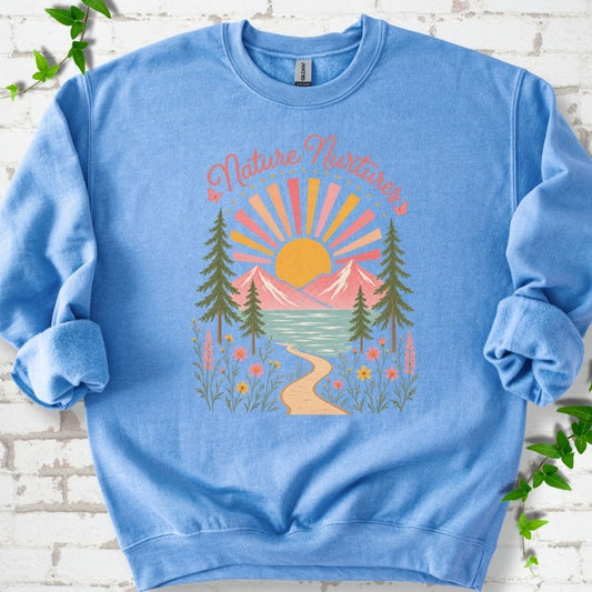 Carolina Blue sweatshirt laid flat, featuring the words 'Nature Nurtures' above a radiating sun, pink mountains, evergreens, blue-green water, a winding path and flowers.