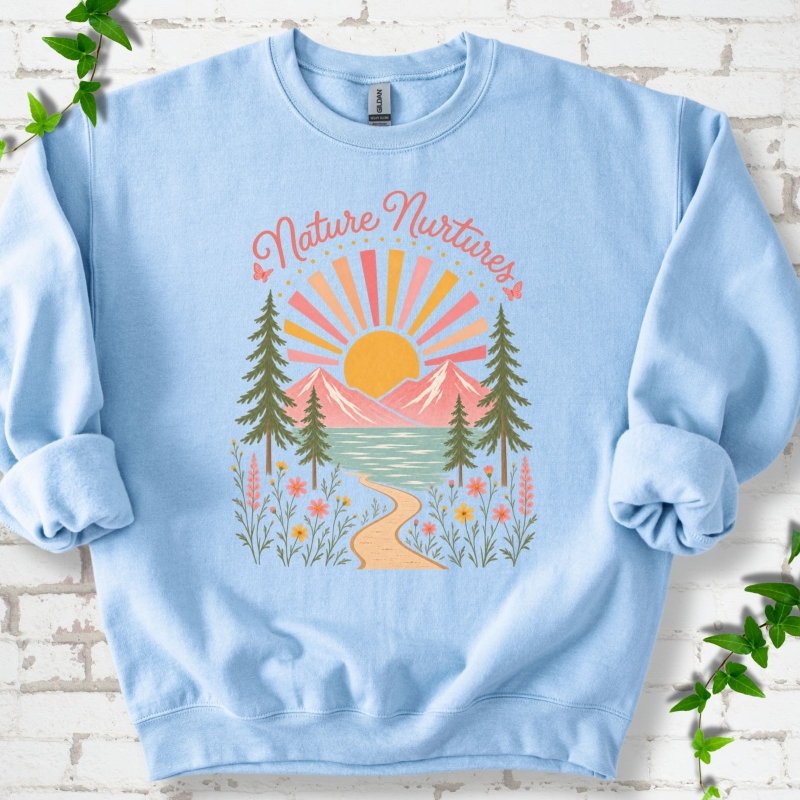 Light Blue sweatshirt laid flat, featuring the words 'Nature Nurtures' above a radiating sun, pink mountains, evergreens, blue-green water, a winding path and flowers.
