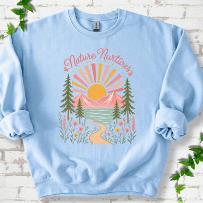 Light Blue sweatshirt laid flat, featuring the words 'Nature Nurtures' above a radiating sun, pink mountains, evergreens, blue-green water, a winding path and flowers.