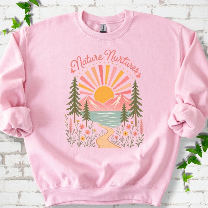 Light Pink sweatshirt laid flat, featuring the words 'Nature Nurtures' above a radiating sun, pink mountains, evergreens, blue-green water, a winding path and flowers.