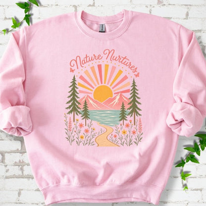 Light Pink sweatshirt laid flat, featuring the words 'Nature Nurtures' above a radiating sun, pink mountains, evergreens, blue-green water, a winding path and flowers.