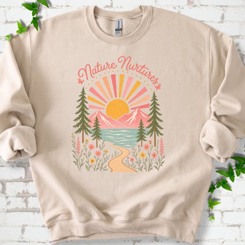 Sand sweatshirt laid flat, featuring the words 'Nature Nurtures' above a radiating sun, pink mountains, evergreens, blue-green water, a winding path and flowers.
