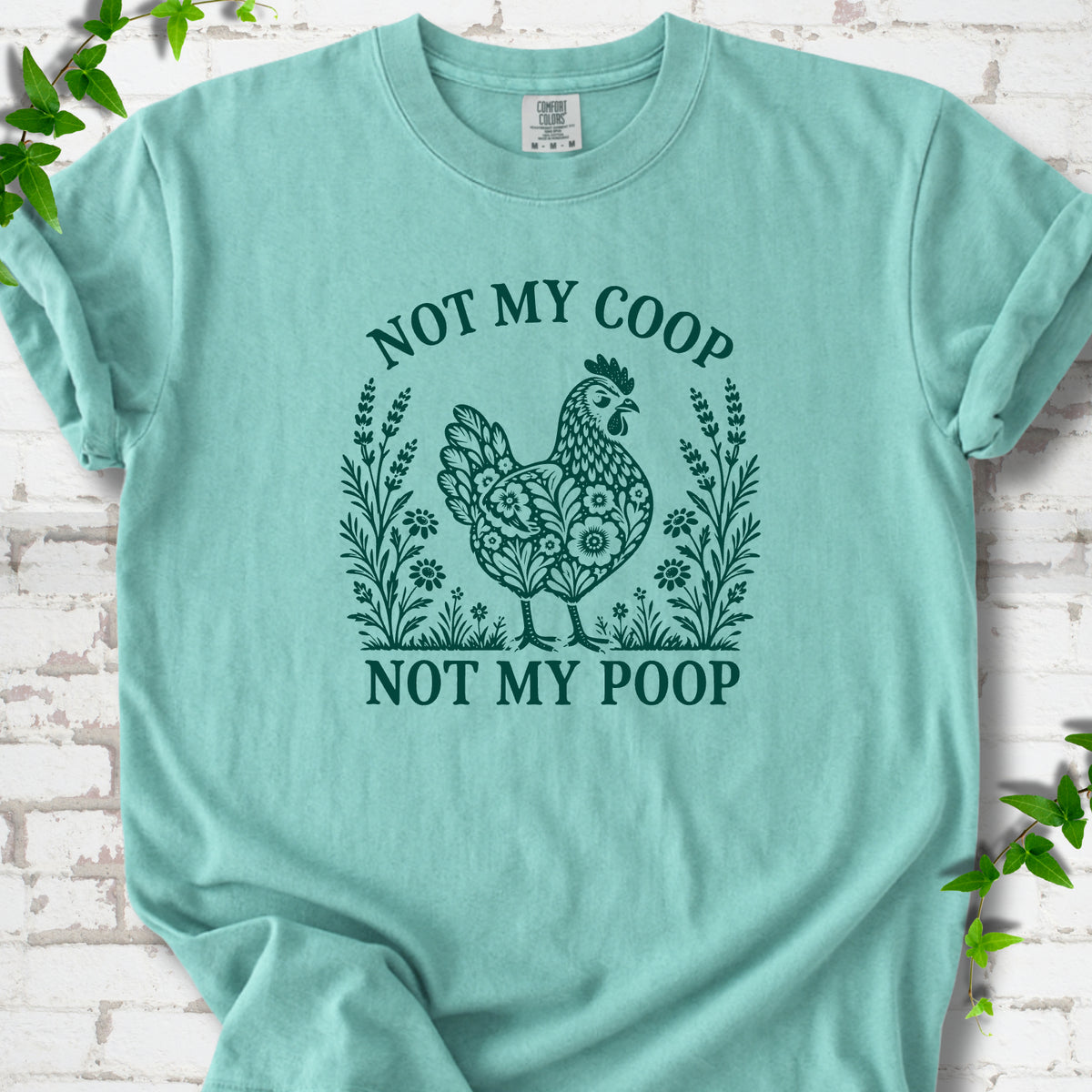 Chalky Mint T-shirt flat lay with a deep teal folk-art hen surrounded by wildflowers and lavender, "Not my coop" arched above and "Not my poop" below, on white brick with ivy.