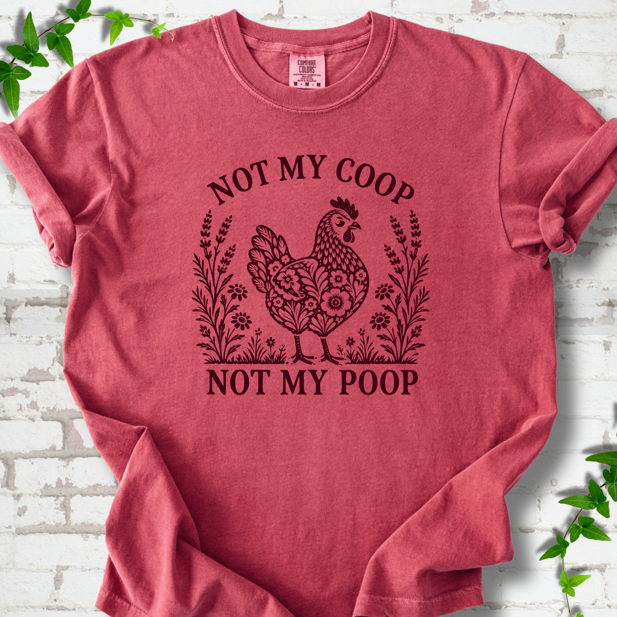 Crimson T-shirt flat lay with a dark burgundy folk-art hen surrounded by wildflowers and lavender, "Not my coop" arched above and "Not my poop" below, on white brick with ivy.