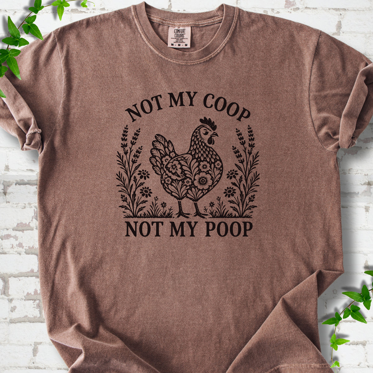 Espresso T-shirt flat lay with a dark brown folk-art hen surrounded by wildflowers and lavender, "Not my coop" arched above and "Not my poop" below, on white brick with ivy.