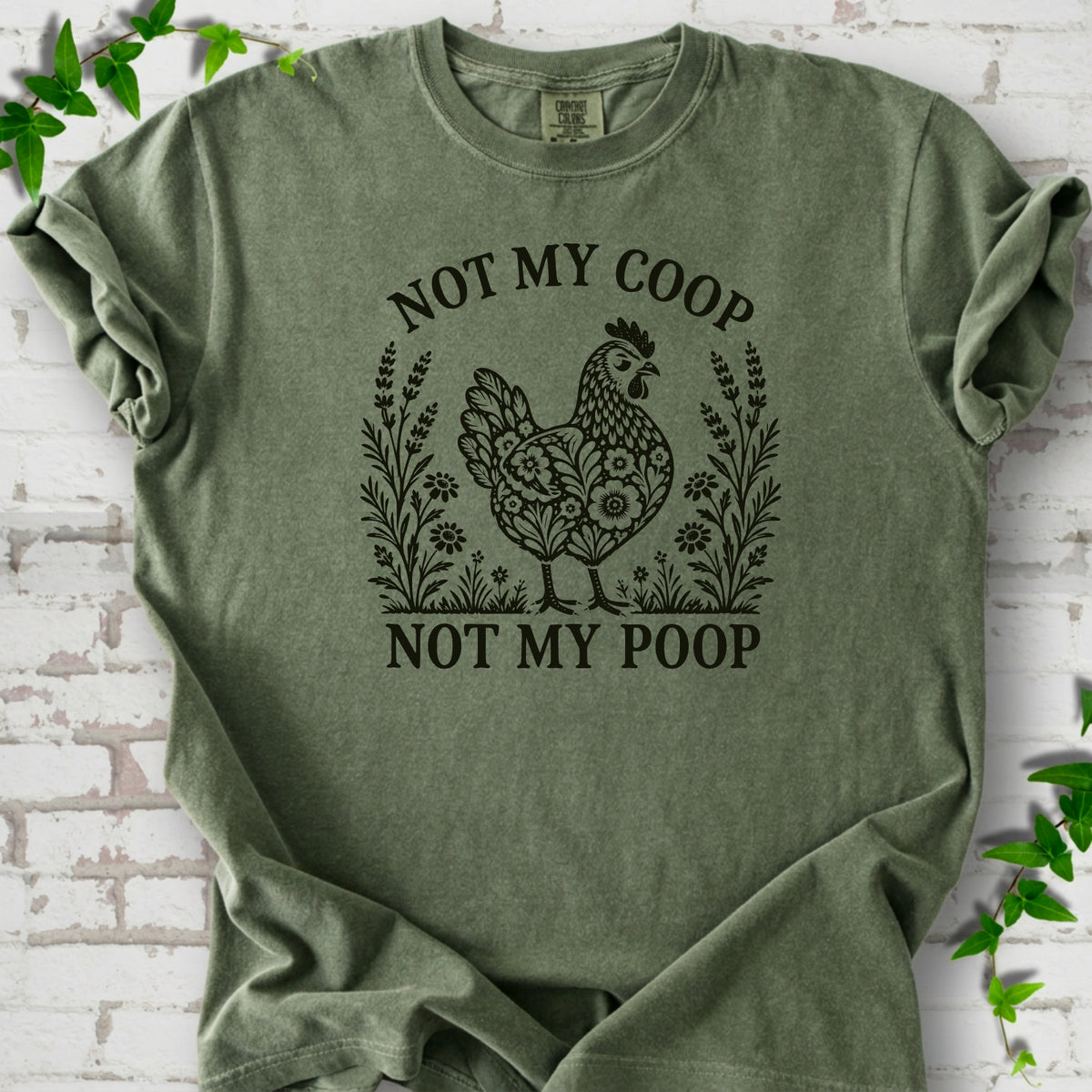 Moss T-shirt flat lay with a near-black olive folk-art hen surrounded by wildflowers and lavender, "Not my coop" arched above and "Not my poop" below, on white brick with ivy.