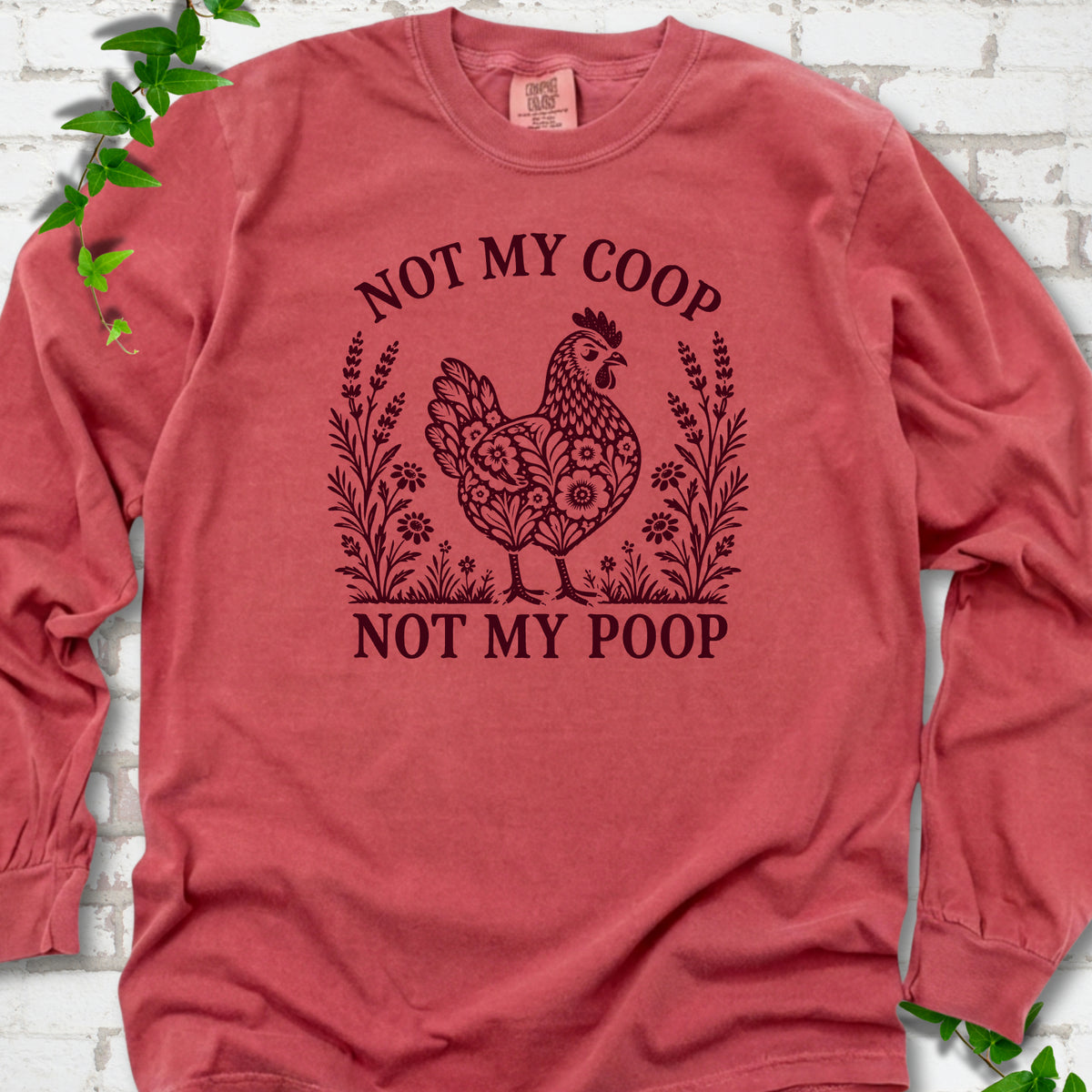 Crimson long sleeve T-shirt flat lay with a dark burgundy folk-art hen surrounded by wildflowers and lavender, "Not my coop" arched above and "Not my poop" below, on white brick with ivy.
