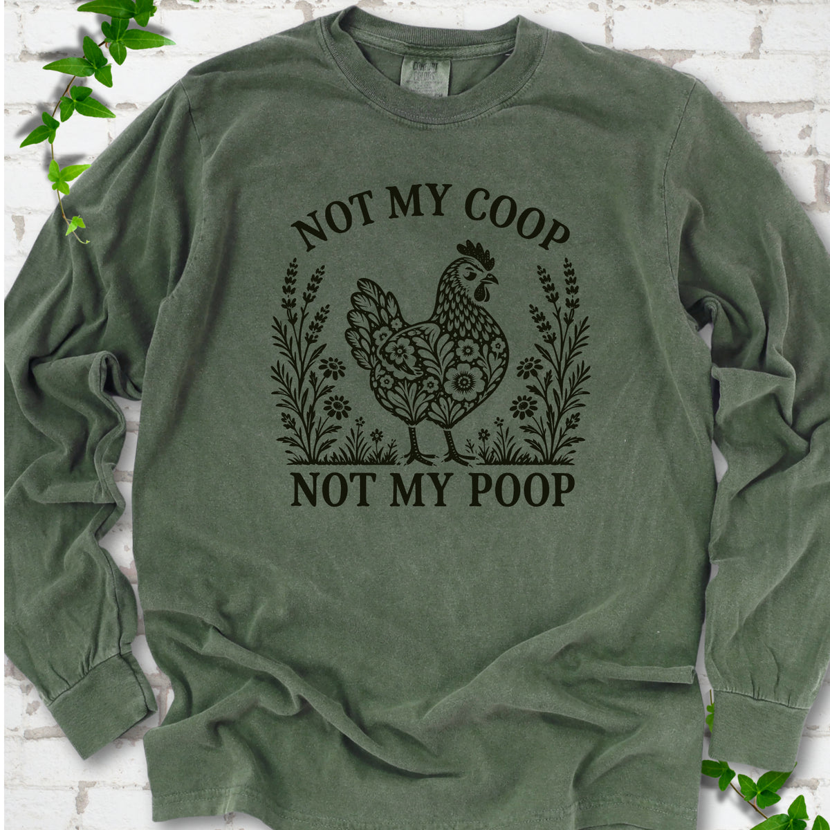 Hemp long sleeve T-shirt flat lay with a near-black olive folk-art hen surrounded by wildflowers and lavender, "Not my coop" arched above and "Not my poop" below, on white brick with ivy.