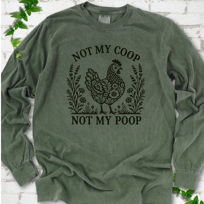 Hemp long sleeve T-shirt flat lay with a near-black olive folk-art hen surrounded by wildflowers and lavender, "Not my coop" arched above and "Not my poop" below, on white brick with ivy.