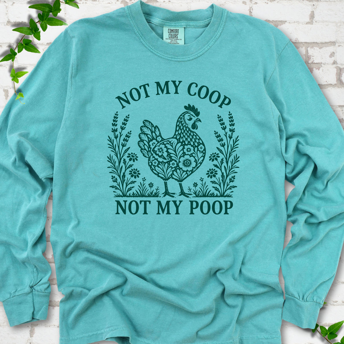 Seafoam long sleeve T-shirt flat lay with a deep teal folk-art hen surrounded by wildflowers and lavender, "Not my coop" arched above and "Not my poop" below, on white brick with ivy.