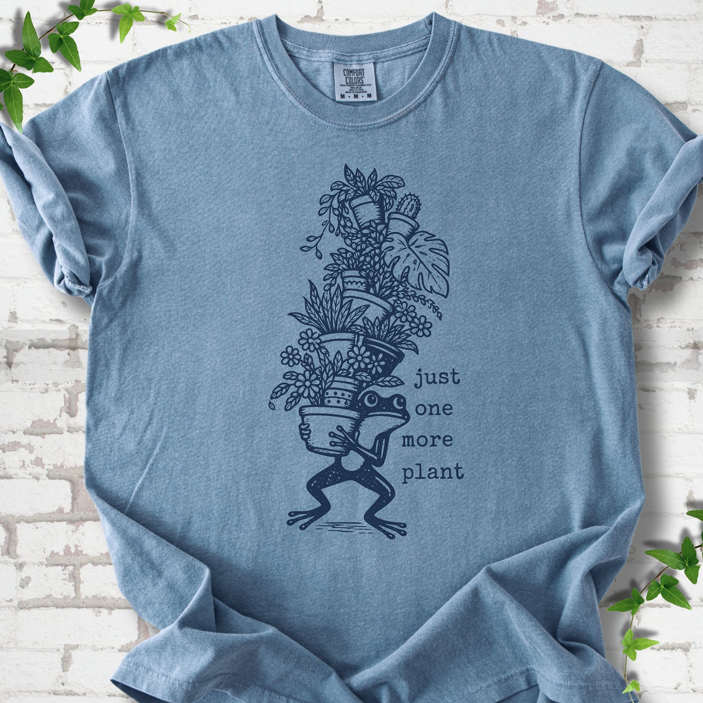 Blue Jean Comfort Colors® T-shirt flat lay featuring a frog carrying a tall stack of houseplants and the words just one more plant.