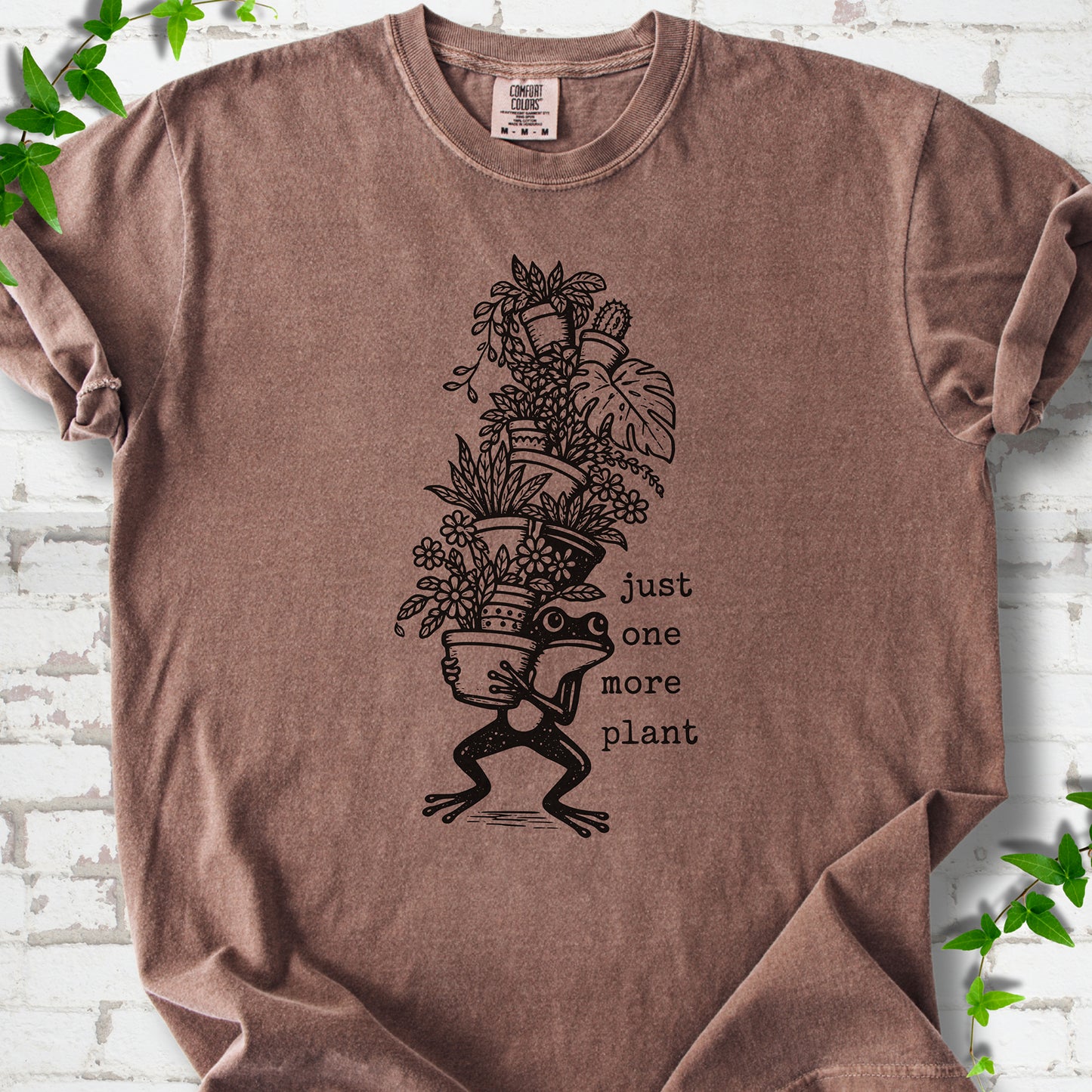 Espresso Comfort Colors® T-shirt flat lay featuring a frog carrying a tall stack of houseplants and the words just one more plant.