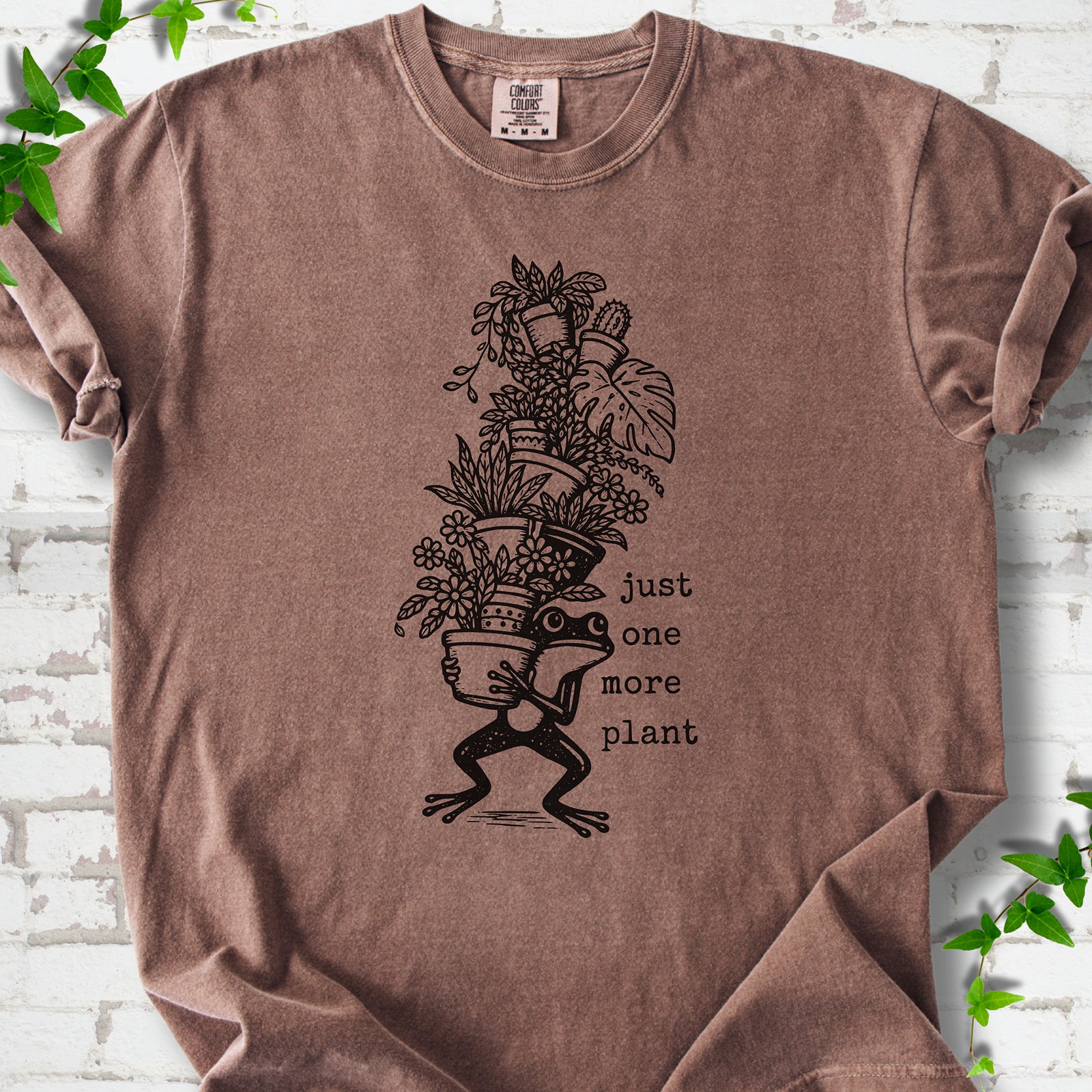 Espresso Comfort Colors® T-shirt flat lay featuring a frog carrying a tall stack of houseplants and the words just one more plant.