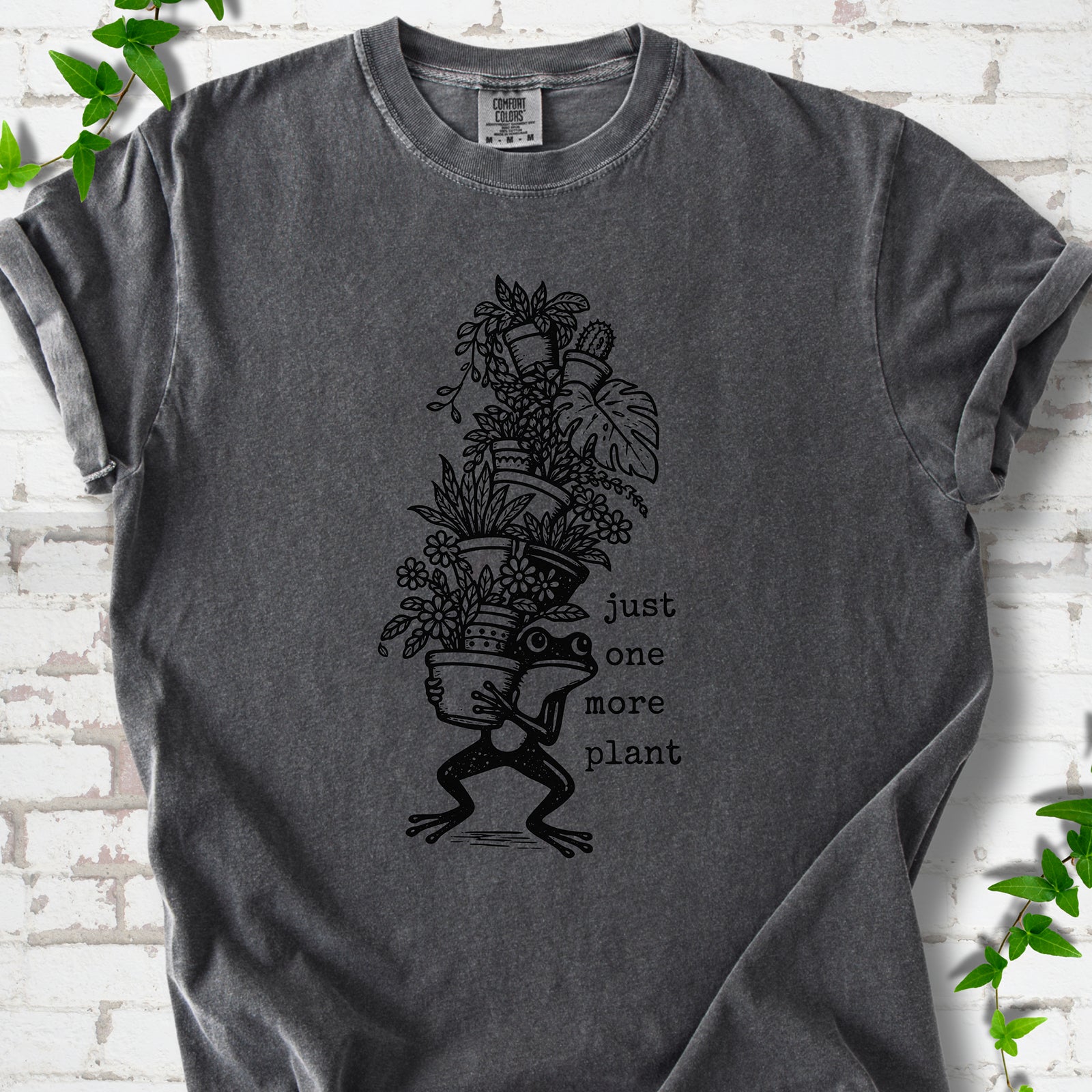 Pepper Comfort Colors® T-shirt flat lay featuring a frog carrying a tall stack of houseplants and the words just one more plant.